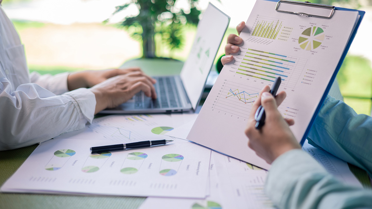 Two professionals analyze charts and graphs on printed reports with a laptop in the background.
