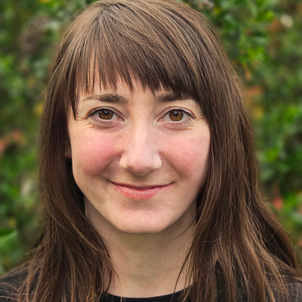 Close-up of a smiling woman with brown hair, outdoors with greenery in the background.
